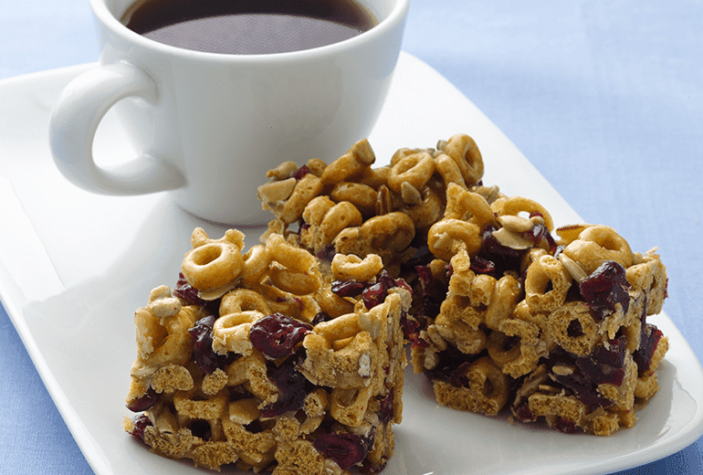 Three bars made from Banana Nut Cheerios, peanut butter, almonds, and cranberries on a plate with a cup of coffee.