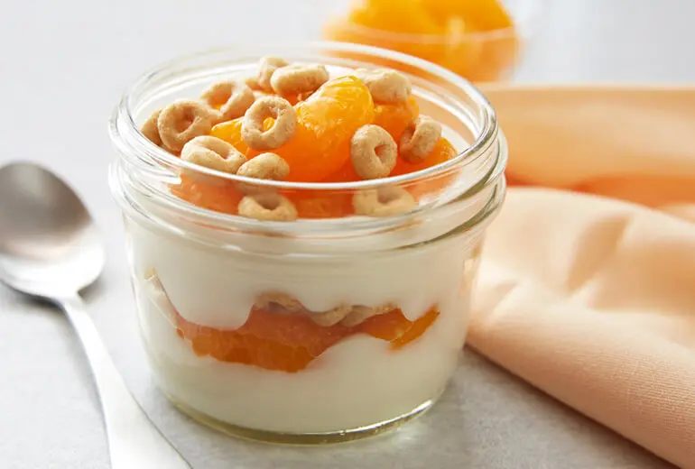 Layers of vanilla yogurt, sweet mandarin oranges and Honey Nut Cheerios in a glass ramekin with a spoon beside it.