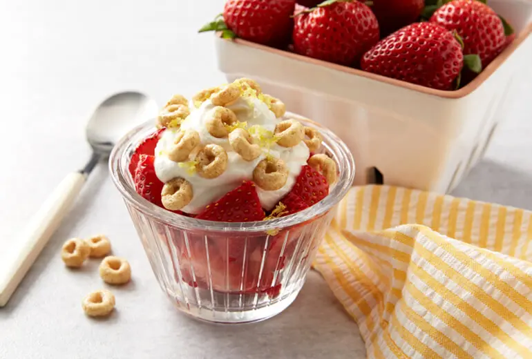 Fresh strawberries topped with vanilla yogurt and Honey Nut Cheerios in a glass ramekin. Fresh strawberries are behind in a basket.
