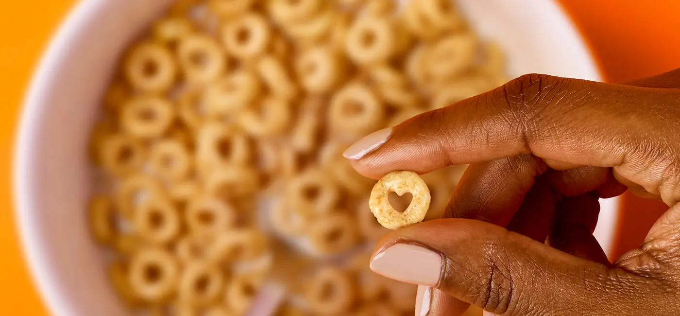 Hand holding a Cheerio with a heart-shaped hole and a bowl of Cheerios in the background.