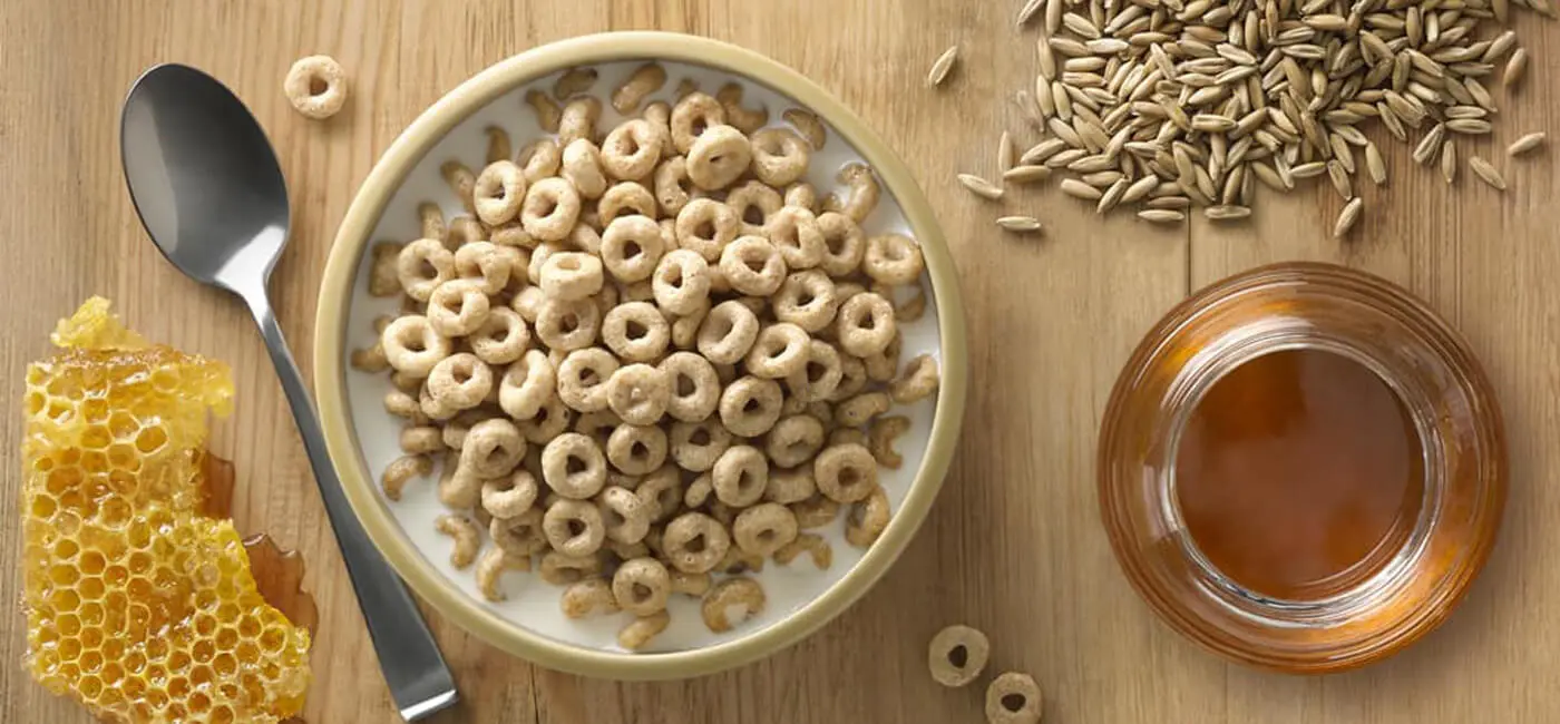 Overhead view of a bowl of Cheerios surrounded by a piece of honeycomb, a pile of grain, and a jar of honey.