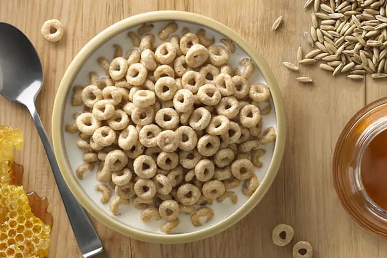 Overhead view of a bowl of Cheerios surrounded by a piece of honeycomb, a pile of grain, and a jar of honey.