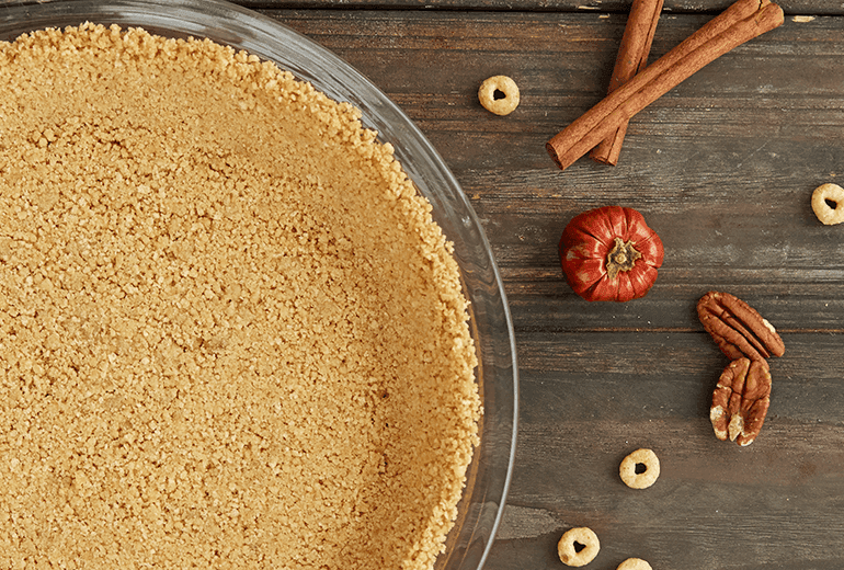 Overhead view of gluten free Cheerios crumb crust with pecans, pumpkin spice, and cinnamon pressed into a glass pie plate.