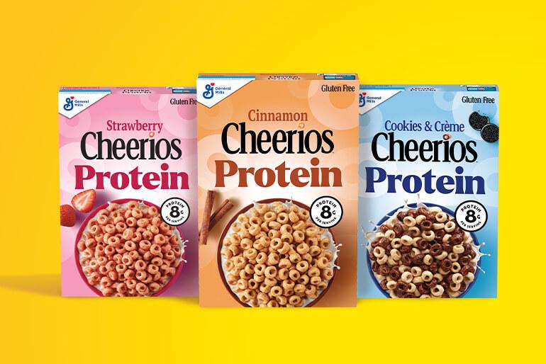 Boxes of Strawberry Cheerios Protein, Cheerios Cookies & Creme and Cinnamon Cheerios Protein are displayed against a yellow background.