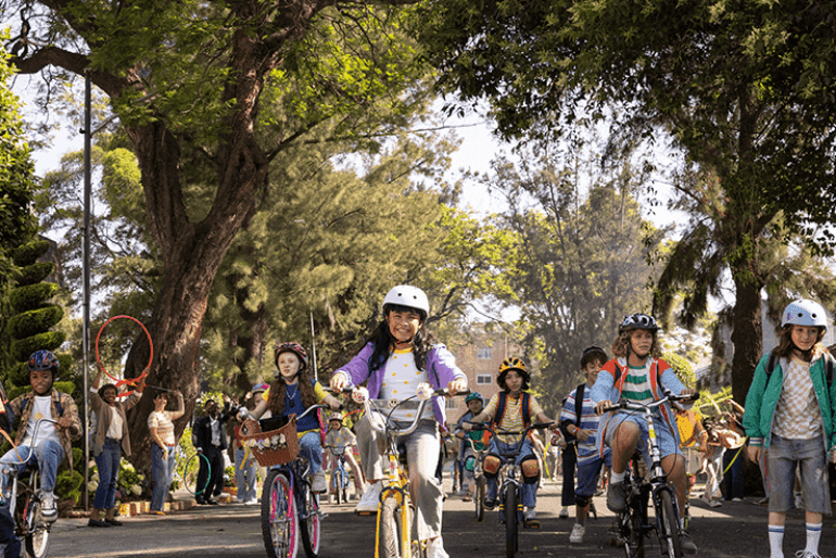 A group of children riding bicycles and skateboards down a street lined with trees and people. Some children wear helmets for safety. In the background, adults stand near the sidewalk, some holding hula hoops and others watching the children.