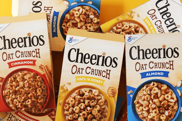 Cuatro cajas de Cheerios Oat Crunch encima de otras cajas, con las cajas exteriores con sabor a almendra y las dos interiores con sabor a canela y avena con miel.