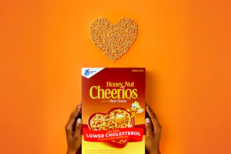 Una caja de Honey Nut Cheerios es sostenida por los lados con las manos para no cubrir el logo; encima de la caja hay un corazón hecho de cereal Honey Nut Cheerios.