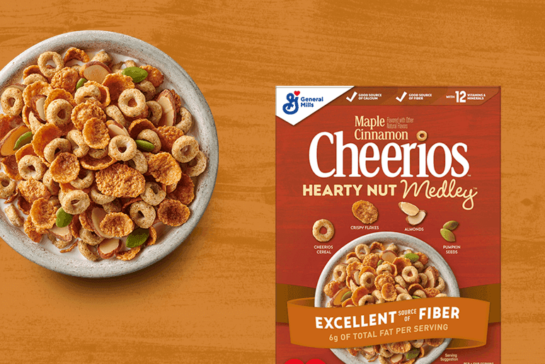 Un tazón de Cheerios Hearty Nut Medley Maple Cinnamon con leche, al lado de la caja de la que provienen. Todo está sobre una tabla de madera.