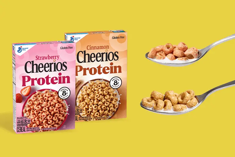 Las cajas de proteína Cheerios de fresa y proteína Cheerios de canela se muestran sobre un fondo amarillo.