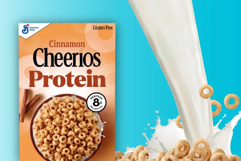 A box of Cinnamon Cheerios Protein cereal is set against a bright blue background with a splash of milk and flying cereal pieces to the right. The orange box features the product name prominently, along with cinnamon sticks and a bowl of cereal at the bottom. Labels highlight "Gluten Free" and "8g Protein with Milk," emphasizing its nutritional benefits.