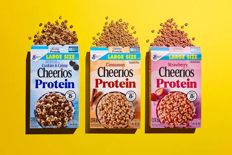Three boxes of Cheerios Protein cereal—Cookies & Crème, Cinnamon, and Strawberry—are displayed side by side on a bright yellow background. Each box is labeled "LARGE SIZE" and highlights "8g Protein" and "Gluten Free." Cereal pieces spill out from the tops of the boxes in a scattered pattern, emphasizing variety and energy.