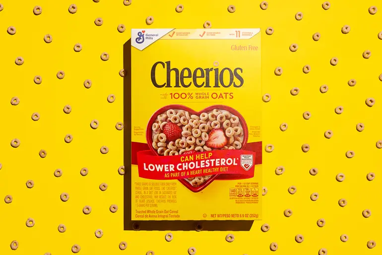 A box of original Cheerios cereal is centered against a bright yellow background patterned with scattered Cheerios pieces. The box features the Cheerios logo, "100% Whole Grain Oats," and a red heart filled with cereal and a strawberry, promoting heart health and cholesterol reduction. It also notes the cereal is gluten-free and displays the General Mills logo in the top left.