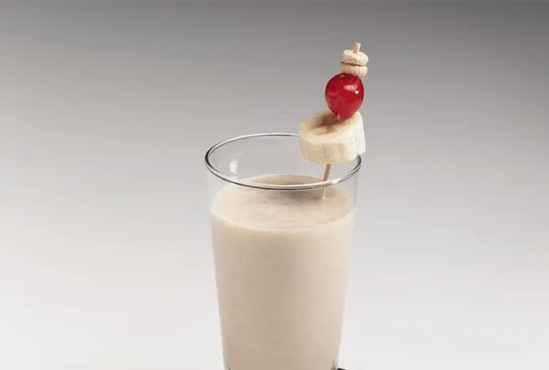 Cereal-Milk Shake with a slice of banana, a glazed cherry and some cheerios all on a skewer sticking out of it.