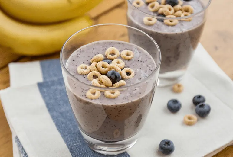 Two Blueberry-Banana Cheerios Cereal Smoothies in glasses with bananas, cheerios and blueberries scattered about them.