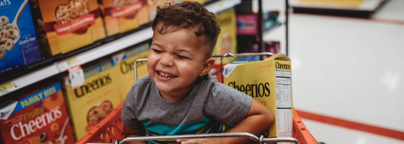 A grinning toddler boy and a box of Cheerios Oats Cereal together in a grocery shopping cart in the cereal aisle.