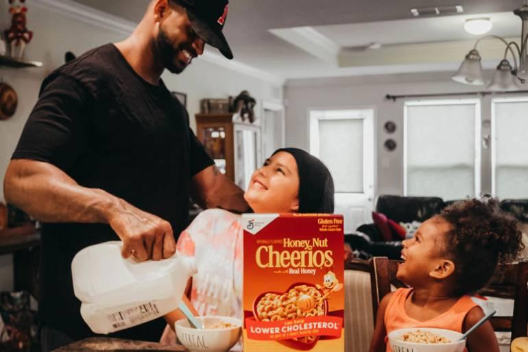A smiling father pouring milk into a bowl of Honey Nut Cheerios cereal for two happy girls.