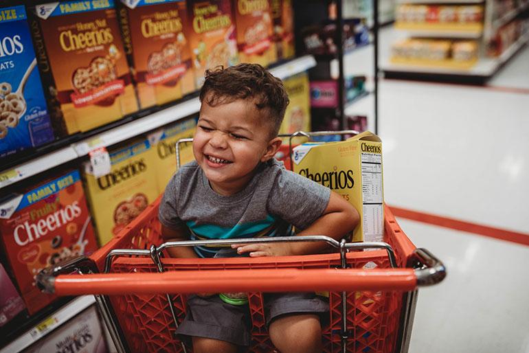 A grinning toddler boy and a box of Cheerios Oats Cereal together in a grocery shopping cart in the cereal aisle.