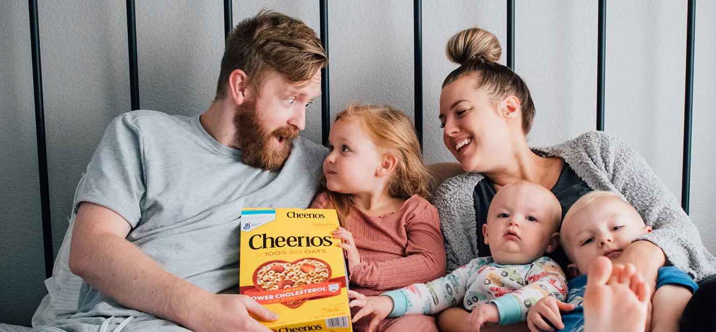 Parents relax with their 3 young children, the dad and eldest daughter are holding a box of Gluten Free Cheerios.