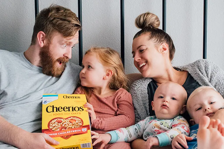 Parents relax with their 3 young children, the dad and eldest daughter are holding a box of Gluten Free Cheerios.