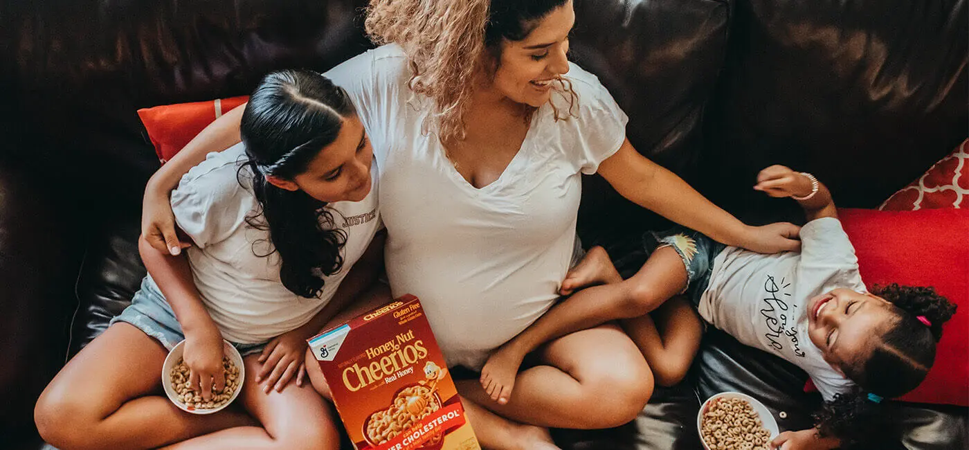 A pregnant mother and her two daughters playing on the couch while the older daughter eats from a bowl of Honey Nut Cheerios.