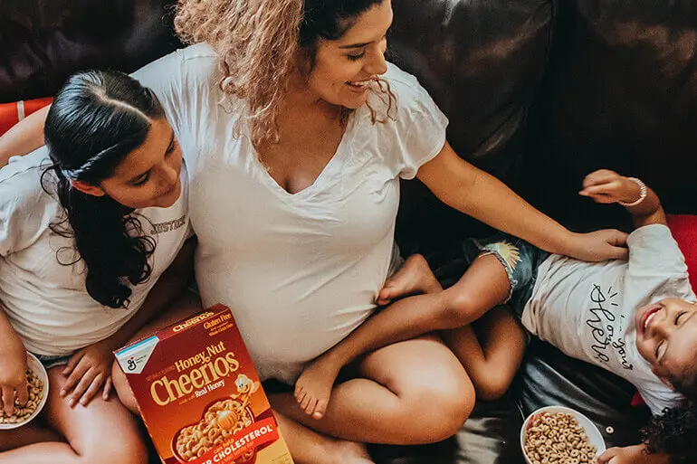 A pregnant mother and her two daughters playing on the couch while the older daughter eats from a bowl of Honey Nut Cheerios.